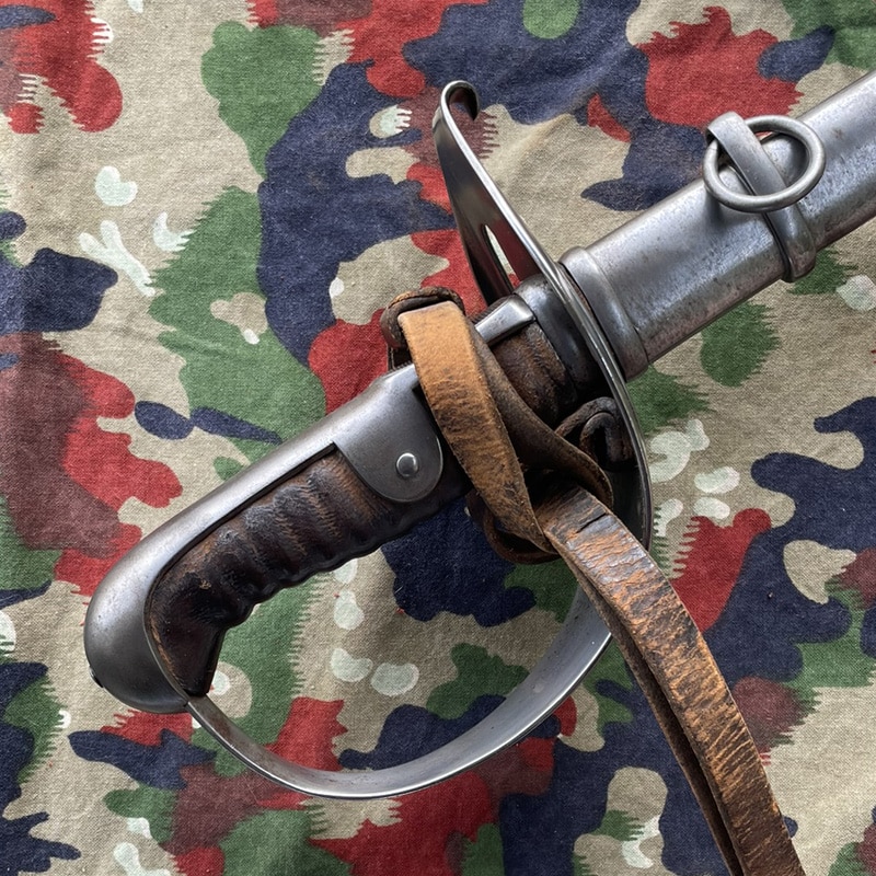 Säbel Ord. 1896 für Mannschaften Waffenfabrik Neuhausen | Fabulafero - Antiquitäten und Einzigartiges Close-up of a steel dagger with a ribbed leather grip and metal guard, strapped with brown leather on a camo fabric background